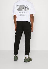 Vit grafisk t-shirt med svart text på ryggen, parat med svarta cargo-byxor med sidofickor och gröna sportskor.