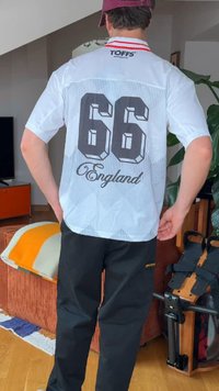Person wearing a white sports jersey with "66" and "England" on the back, paired with black pants and a red cap, indoors.