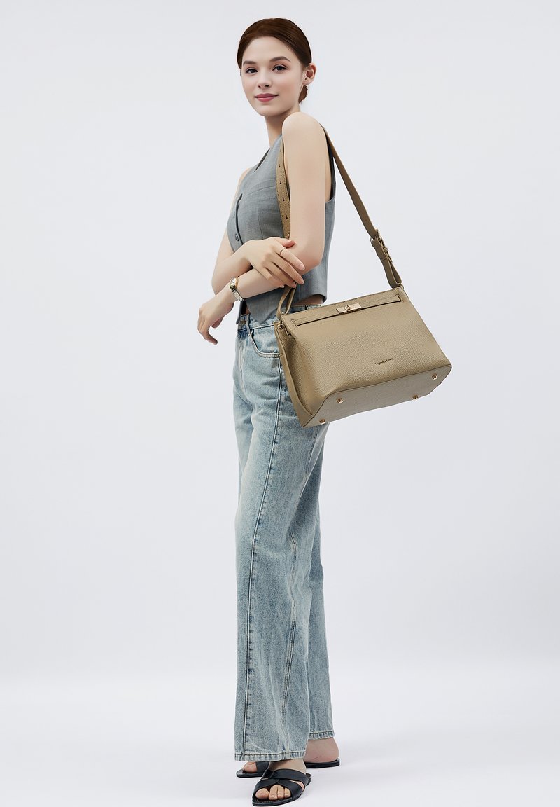 Beige Lederhandtasche mit strukturierter Form, oberen Reißverschluss, goldenen Hardware-Akzenten und vier metallischen Füßen. Mit verstellbarem Riemen über der Schulter getragen.