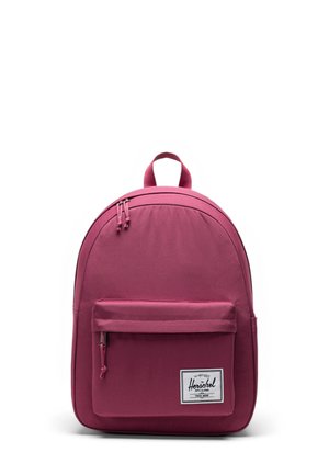 Pink backpack with a smooth fabric texture, rounded shape, top handle, front zip pocket, and a white brand label.