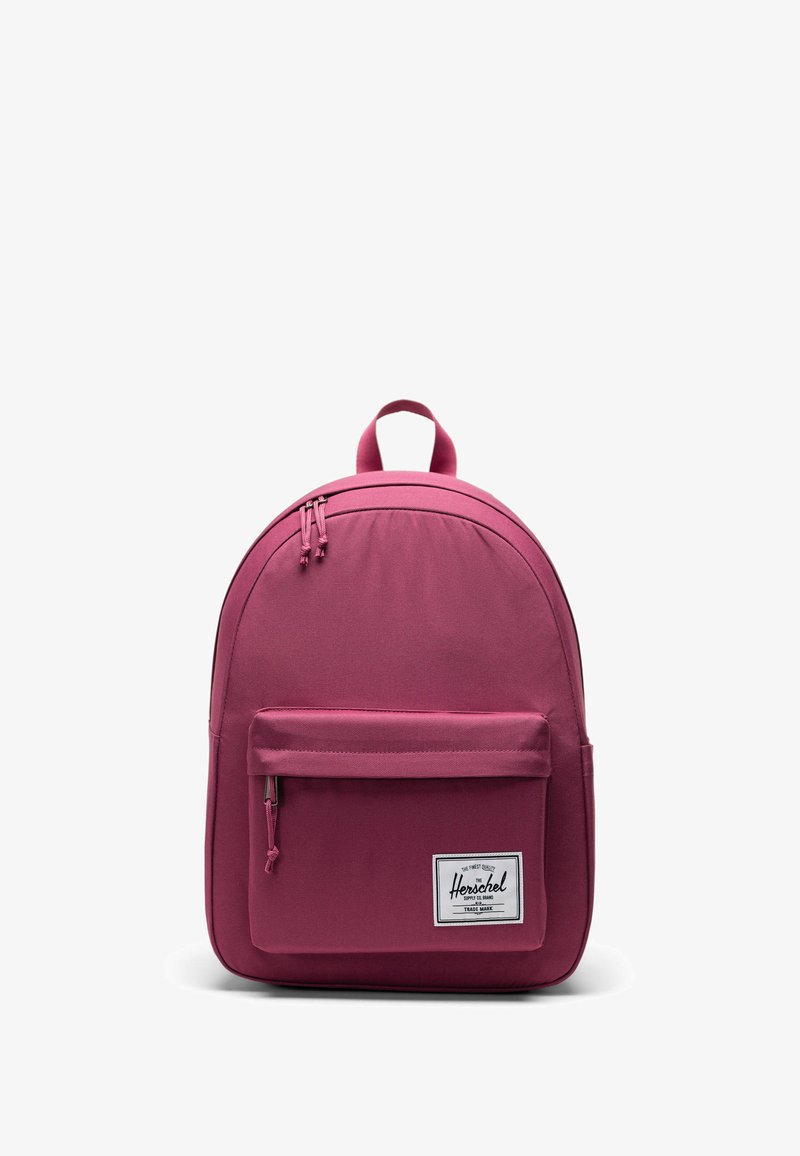 Rosa ryggsäck med en slät tygkvalitet, rund form, handtag upptill, framficka med dragkedja och en vit varumärkesetikett.