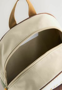 Beige ryggsäck med bruna detaljer och tan-handtag. Har en smidig dragkedja och ett rymligt inre foder i en enfärgad tyg.