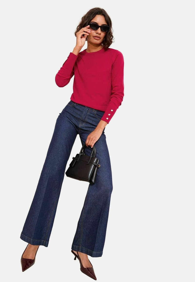 Top roșu cu mâneci lungi, accentuat cu nasturi, purtat cu blugi negri, largi. Modelul ține o geantă mică neagră și poartă ochelari de soare negri.