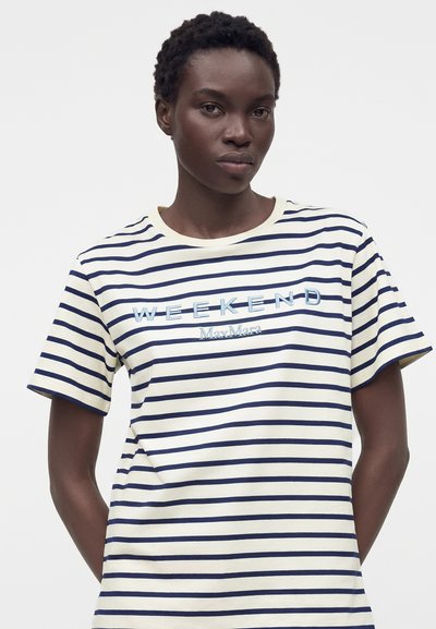 Γυναίκα με κοντά σγουρά μαλλιά που φοράει λευκό και ναυτικό ριγέ μπλουζάκι με το κείμενο "WEEKEND Max Mara" μπροστά.