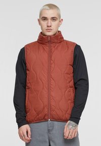Red quilted vest with high collar and front zipper. Worn over a long-sleeve black top. Features a smooth texture and subtle wave pattern.
