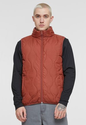 Chaleco acolchado rojo con cuello alto y cremallera frontal. Lleva puesto sobre una camiseta negra de manga larga. Presenta una textura suave y un sutil patrón de olas.