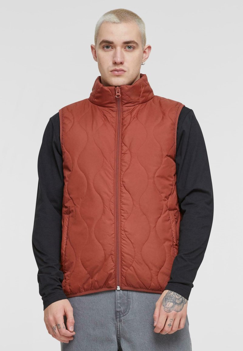 Red quilted vest with high collar and front zipper. Worn over a long-sleeve black top. Features a smooth texture and subtle wave pattern.