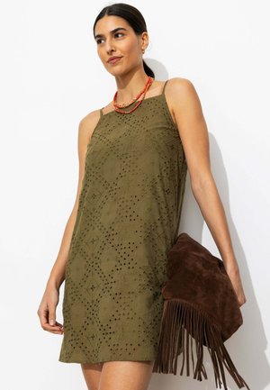 Femme portant une robe sans manches olive à œillets, tenant une pochette en daim marron à franges, et portant des colliers orange superposés.