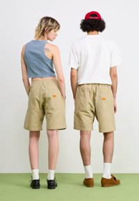 Beige cargo shorts with an elastic waistband and two back pockets. A cropped blue tank top and a white t-shirt are worn. Green floor background.