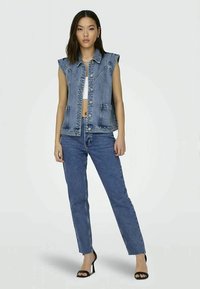 Džinsinis be rankovių švarkas su sagų priekiu, krūtinės kišenėmis ir struktūrizuotu dizainu, derinamas su aukštu liemeniu ir tiesiomis mėlynomis džinsinėmis kelnėmis.