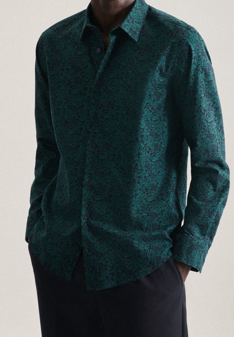 Camicetta a maniche lunghe verde con motivo, dotata di chiusura a bottoni e colletto. Presenta un design floreale testurizzato su uno sfondo scuro.