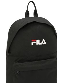 Sac à dos en tissu noir avec un dessus arrondi, comportant une poche avant et un logo FILA rouge et blanc. Poignée de transport robuste attachée.