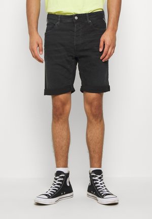 Mann trägt hochgekrempelte schwarze Jeansshorts, weiße Socken, schwarze High-Top-Sneaker und ein hellgelbes Hemd und steht vor einem schlichten weißen Hintergrund.