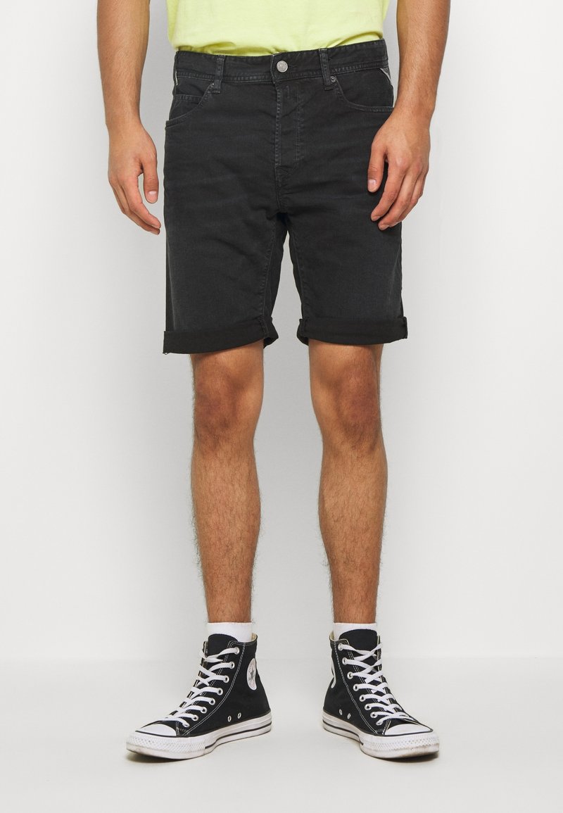 Homme portant un short en jean noir retroussé, des chaussettes blanches, des baskets montantes noires et une chemise jaune clair, debout devant un fond blanc uni.