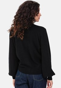 Pull noir en maille avec manches longues légèrement bouffantes, col rond, poignets et ourlet côtelés. Porté avec un jean bleu.
