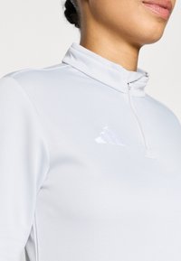 Ανοιχτό γκρι αθλητικό μπλουζάκι με μακριά μανίκια, ψηλό γιακά και φερμουάρ έως το ένα τέταρτο. Διαθέτει διακριτικό αντανακλαστικό λογότυπο Adidas στο στήθος.