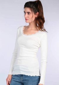 Weißes Langarmoberteil mit einem gewellten Spitzenrundhalsausschnitt, gerippter Textur und eng anliegender Silhouette. Getragen mit blauen Jeans.