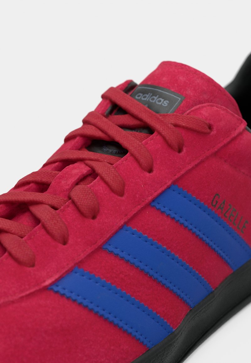 Baskets en daim rouge avec des bandes bleues, des lacets texturés et une semelle noire. Présente le logo Adidas et le marquage « GAZELLE ».
