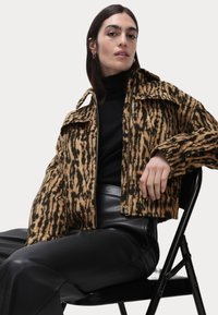 Veste courte à motifs marron et noir avec fermeture éclair à l'avant et deux poches poitrine, associée à un pantalon en cuir noir. Modèle assis sur une chaise.