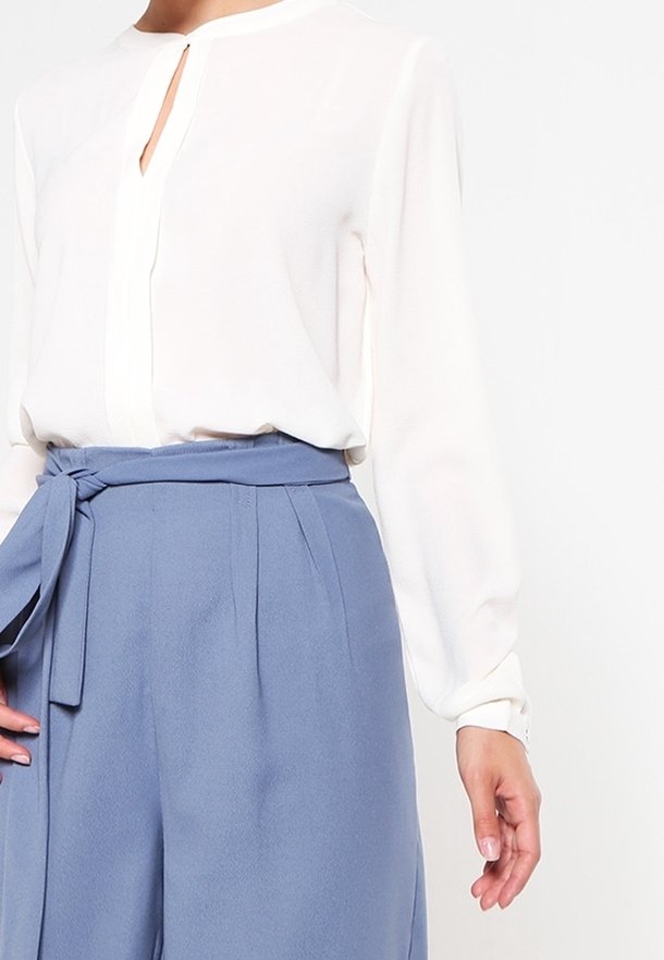 Blouse blanche à manches longues avec une encolure fendue, associée à un pantalon taille haute bleu clair doté d'une ceinture nouée et de plis.