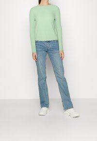 Světle zelený žebrovaný svetřík s dlouhými rukávy a vlnitým lemem, doplněný modrými zvonovými džíny a bílými teniskami.