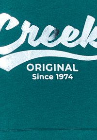 Felpa turchese con un grafico argentato brillante con la scritta "Creek" e testo bianco che recita "ORIGINAL Since 1974". Tessuto morbido con un design casual.