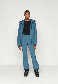 Egy nő áll, teal színű sídzsekiben és nadrágban, fekete pólóval és sícipőben, egyszínű világos háttér előtt.