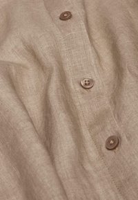Beige Leinenhemd mit strukturierter Oberfläche, ausgestattet mit braunen Knöpfen entlang der Vorderseite und einem dezenten Schimmer.