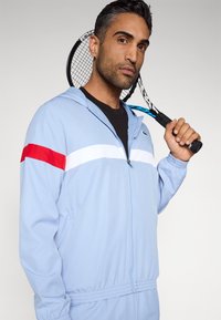 Άνδρας με ανοιχτό μπλε φόρμα κρατώντας μια μαύρη και μπλε ρακέτα τένις ακουμπισμένη στον ώμο του, μπροστά σε ένα απλό φόντο.