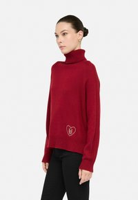 Rød turtleneck sweater i strikket stof med ribbet kant. Har en hjerteapplikation med et design nær bunden til venstre. Modellen bærer sorte bukser.