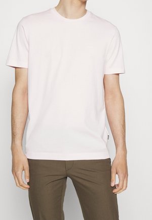 Homme portant un t-shirt à manches courtes col rond rose clair et un pantalon marron, debout devant un fond clair uni.
