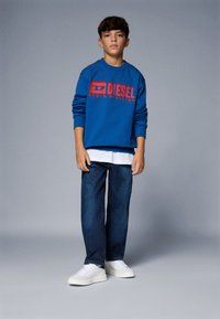 Felpa blu con logo rosso "DIESEL DENIM DIVISION", indossata sopra una maglietta bianca, abbinata a jeans in denim scuro e scarpe da ginnastica bianche.