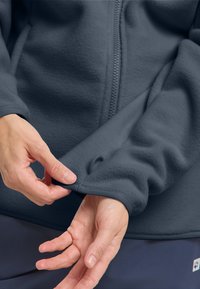 Giacca in fleece grigio scuro con zip, mostra una mano che aggiusta la manica. Il tessuto ha una texture morbida e polsini aderenti per il calore.