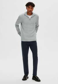 Helles graues Kabelstrickpullover mit einem Halbreißverschluss-Kragen, kombiniert mit marineblauen Hosen und schwarzen Schnürschuhen, die einfarbige und strukturierte Muster aufweisen.