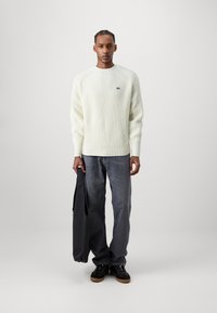 Helles gelbes Rippenpullover mit Rundhalsausschnitt, kombiniert mit dunkelgrauen Jeans. Das Model hält eine schwarze Tragetasche. Schwarze Schuhe mit braunen Sohlen.
