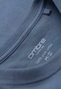 Gros plan sur le col d'un t-shirt bleu montrant l'inscription "Ombre Cotton Life", 100 % coton pur, taille M/48, étiquette imprimée à l'intérieur du col.