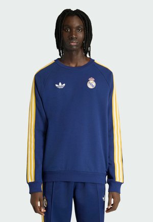 Persona vestida con un conjunto deportivo azul marino con rayas amarillas, que lleva los logotipos de Adidas y Real Madrid, de pie sobre un fondo liso.