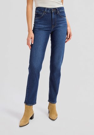 Žena v tmavomodrých džínsach s vysokým pásom a béžových semišových členkových čižmách stojaca na jednoduchom svetlom pozadí.