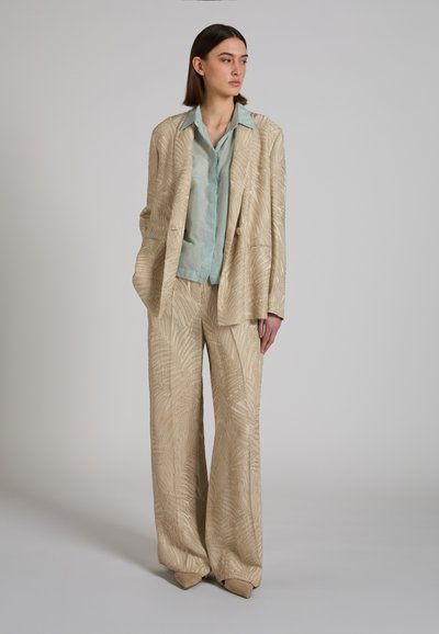 Donna in piedi che indossa un completo a pantaloni larghi beige con fantasia, blazer abbinato sopra una camicia di raso verde pallido e scarpe beige con punta.