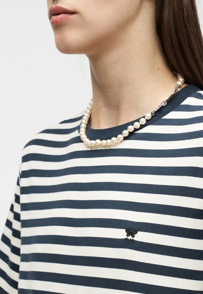 Νεαρό άτομο που φοράει μπλουζάκι με ναυτικό μπλε και άσπρες ρίγες, με ένα μικρό μαύρο λογότυπο και ένα κολιέ με πέρλες και ασημένιο κλείσιμο.
