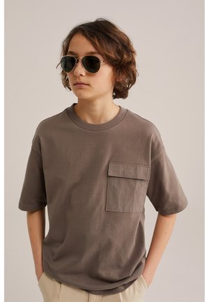 Mladý člověk nosící pilotní sluneční brýle a volné hnědé tričko s velkou kapsou, ruce v kapsách béžových kalhot, neutrální pozadí.