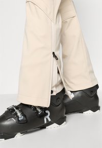 Pantaloni de ski bej cu tivuri cu fermoar, asortați cu bocanci de ski negri cu catarame argintii și un logo R alb, având tălpi cu model.