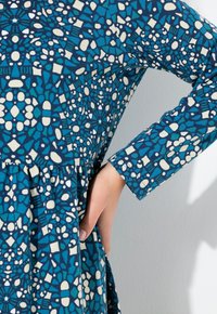 Vestido con estampado azul y blanco, de mangas largas, con una tela texturizada y una cintura fruncida que crea una silueta fluida.