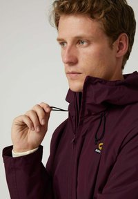 Chaqueta impermeable color burdeos con cuello alto, cordón ajustable y herrajes negros. Presenta un logotipo en el pecho y una textura suave.