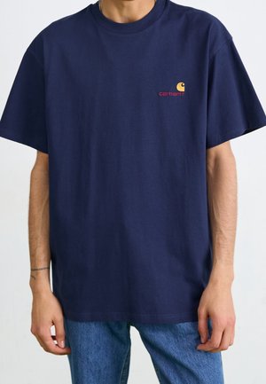 Mężczyzna noszący granatowy t-shirt Carhartt z krótkim rękawem i małym logo na lewej piersi, w zestawieniu z niebieskimi dżinsami.