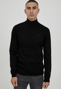 Schwarzer Rollkragenpullover mit langen Ärmeln. Weiches Material, taillierte Passform, gerippter Kragen und Bündchen, dezentes Branding auf der Brust. Trägt man mit grauen Hosen.