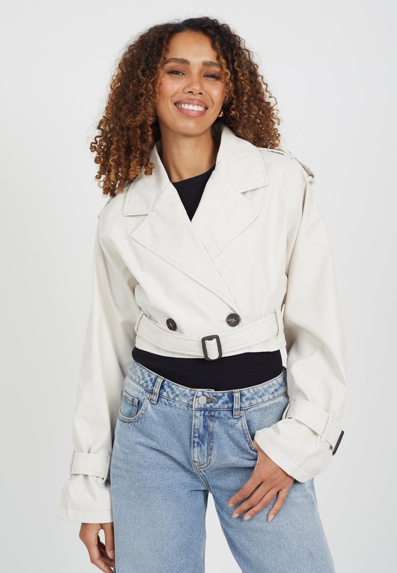 Skórzana beżowa trencz z podwójnym zapięciem na guziki, szerokimi klapami i paskiem w talii; zestawiona z jasnoniebieskimi dżinsami.