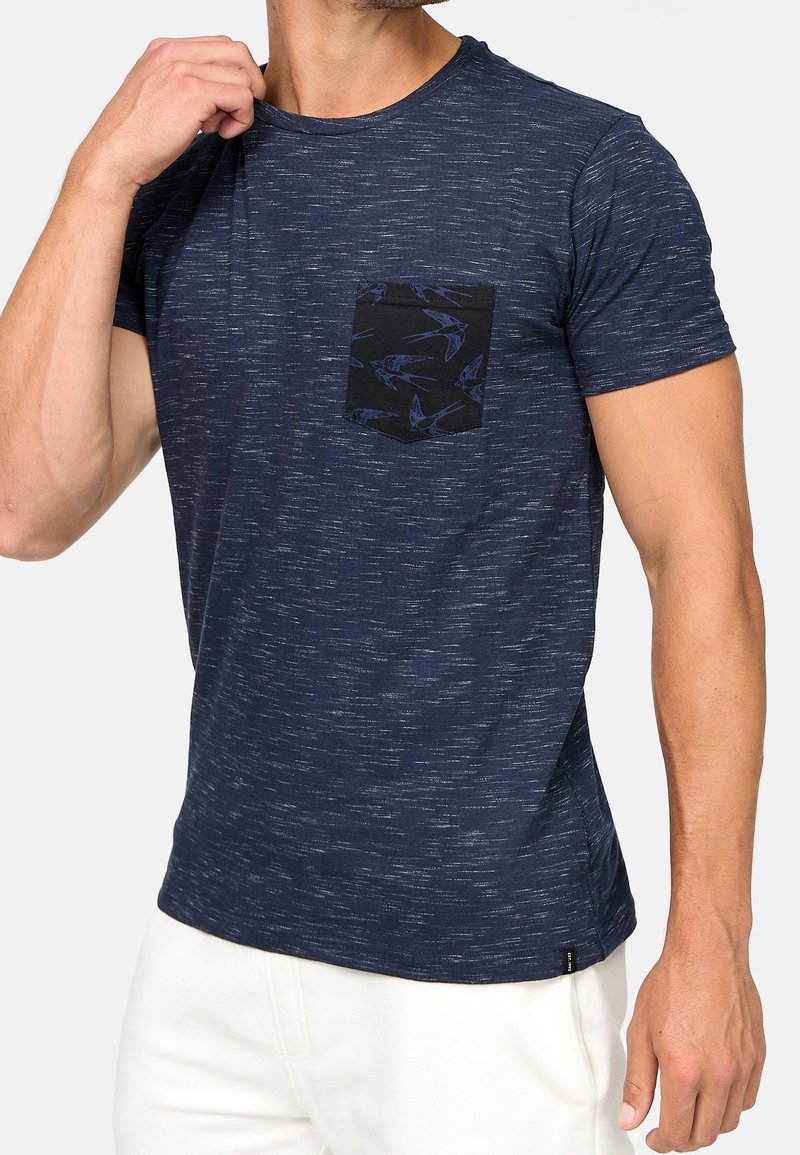 Man wearing a navy blue marled t-shirt with a black chest pocket featuring a blue bird pattern, paired with white pants.