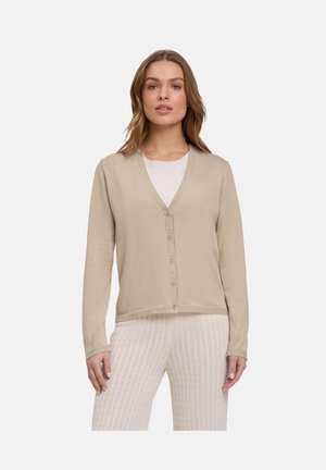 Femme portant un cardigan beige boutonné sur un haut blanc et un pantalon blanc texturé, debout devant un fond clair uni.
