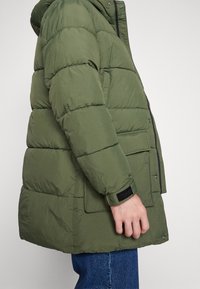 Person iført en olivengrøn polstret jakke med forlommer og en sort lukning på ærmet, kombineret med blå jeans.